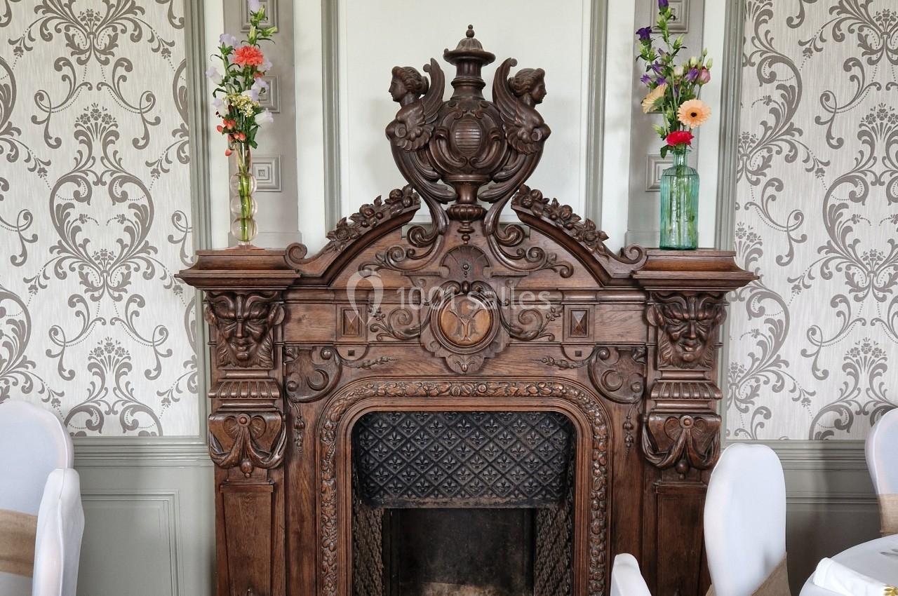 Cheminée en bois sculpté ornée de motifs décoratifs, entourée de chaises blanches et de vases fleuris.