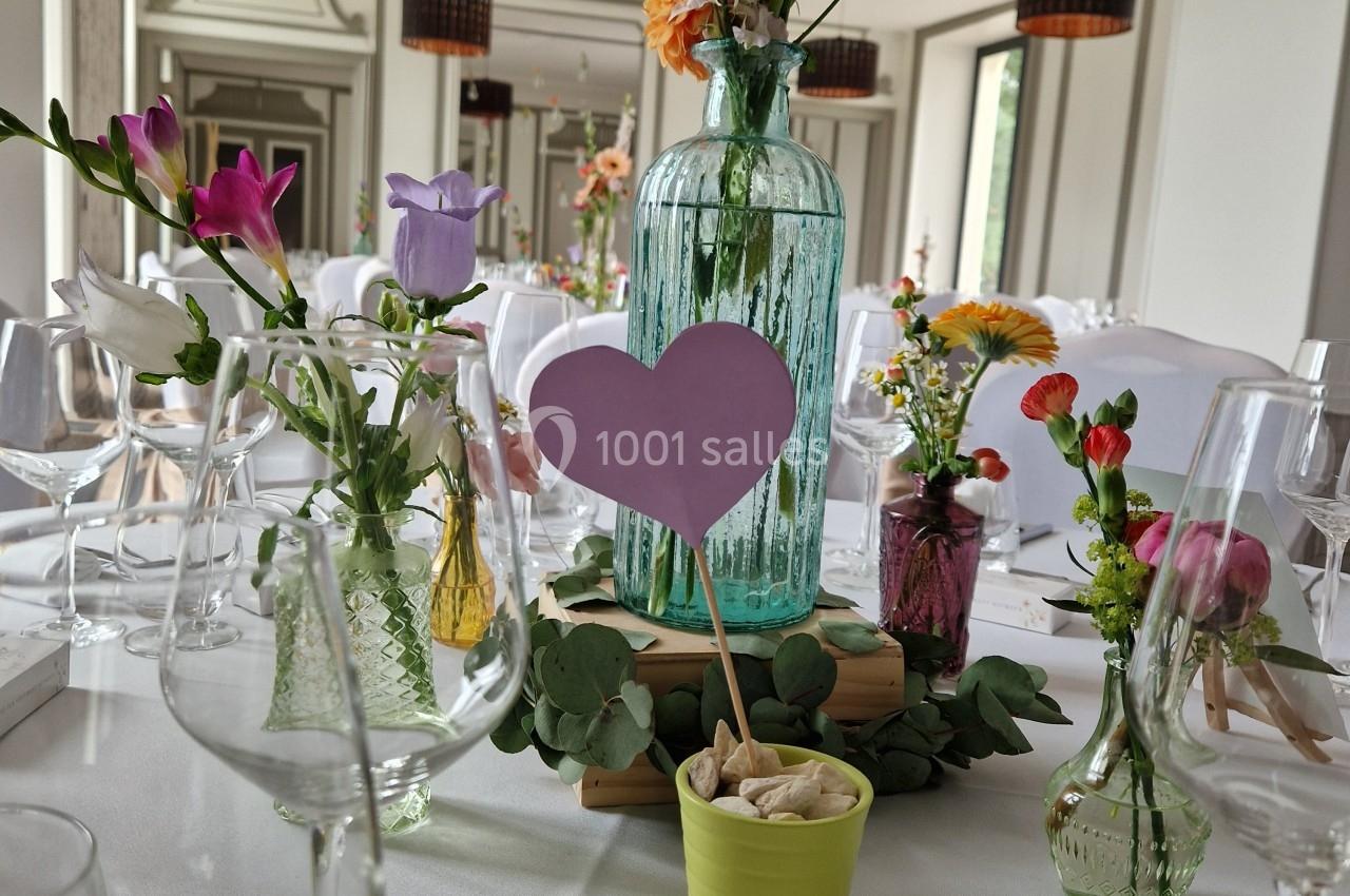 Table décorée pour un événement avec fleurs colorées, vases en verre, et un cœur violet planté dans un pot vert.