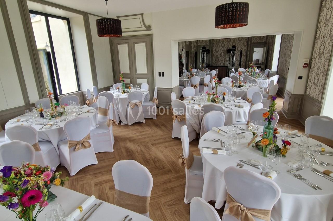 Salle de réception lumineuse avec tables rondes décorées de nappes blanches, fleurs colorées et chaises ornées de nœuds…
