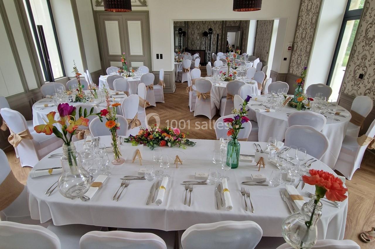 Salle de réception décorée pour un événement, avec tables dressées, nappes blanches et bouquets de fleurs colorées.