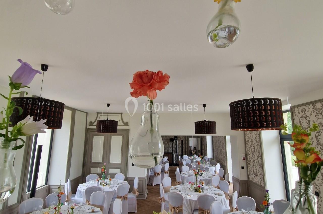 Salle de réception décorée avec des tables dressées et des fleurs suspendues dans des vases en verre.