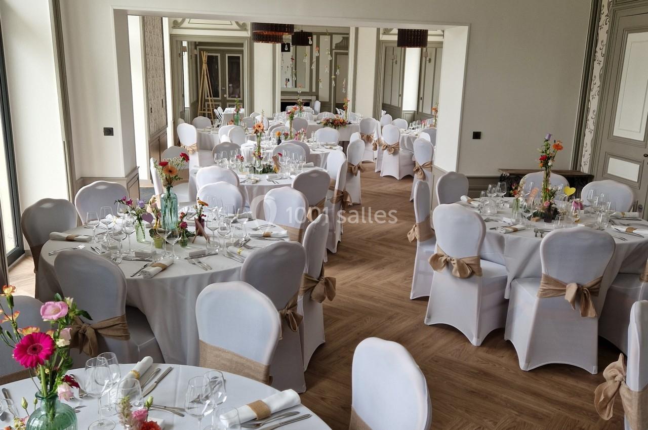 Salle de réception élégante avec tables rondes dressées, nappes blanches et décorations florales colorées.