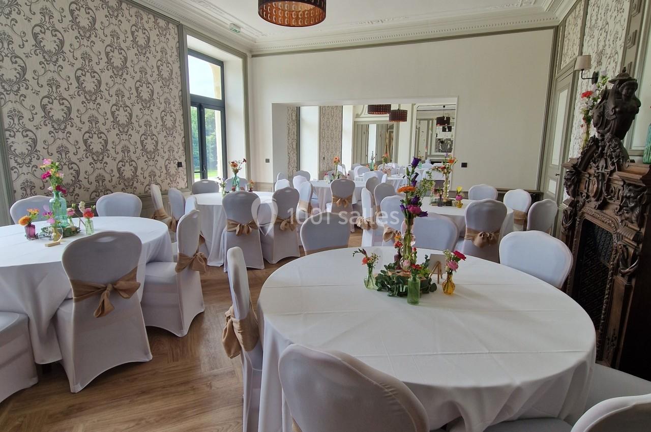 Salle élégante avec tables rondes dressées, nappes blanches, chaises décorées et lumière naturelle entrant par les fenêtres.