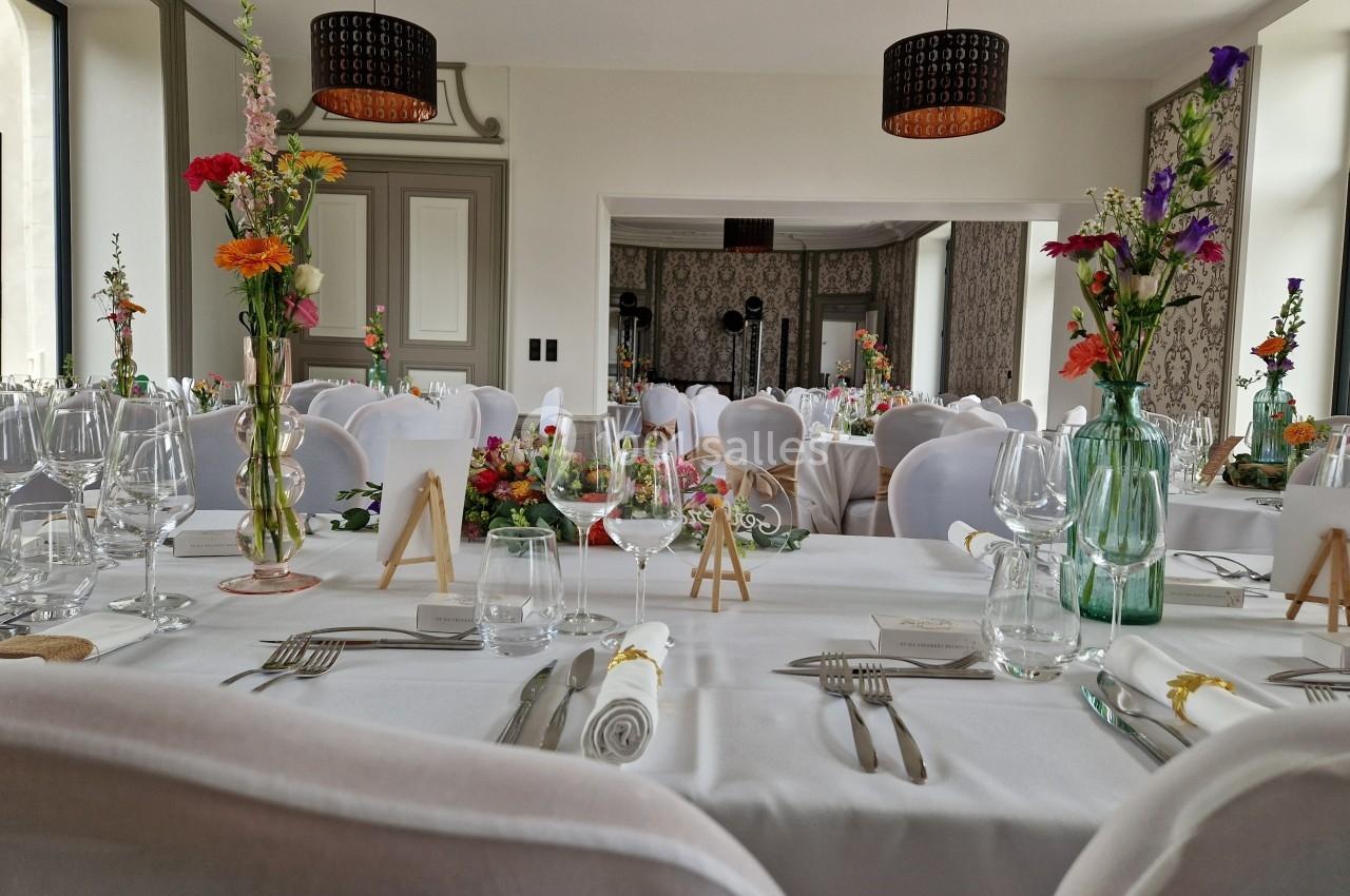 Salle de réception décorée avec des tables dressées, nappes blanches, fleurs colorées et chaises blanches.