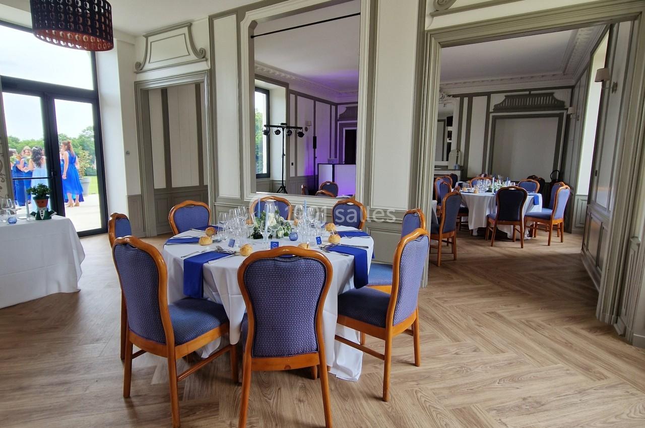 Salle élégante avec tables dressées, chaises bleues et luminaires suspendus, parquet clair et grandes fenêtres.