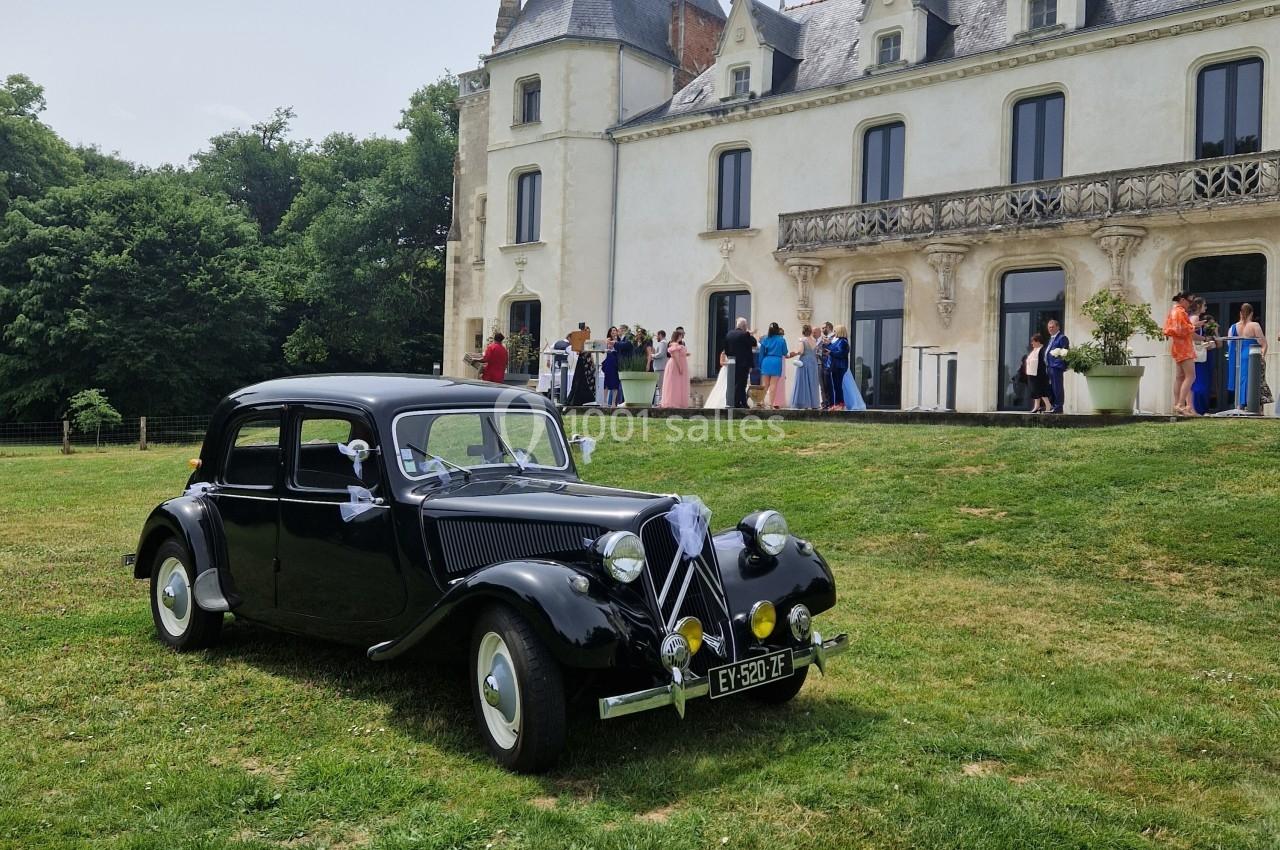 Voiture ancienne noire garée sur une pelouse devant un château, avec des invités en tenue élégante à l'arrière-plan.