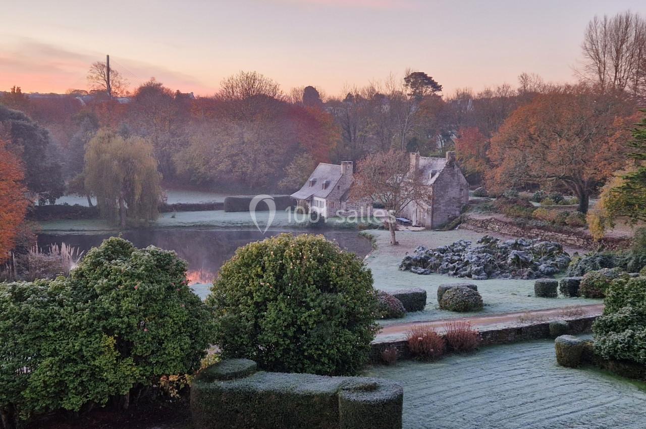 Paysage automnal avec un jardin givreux, un étang reflétant le ciel et deux maisons en pierre entourées d'arbres.