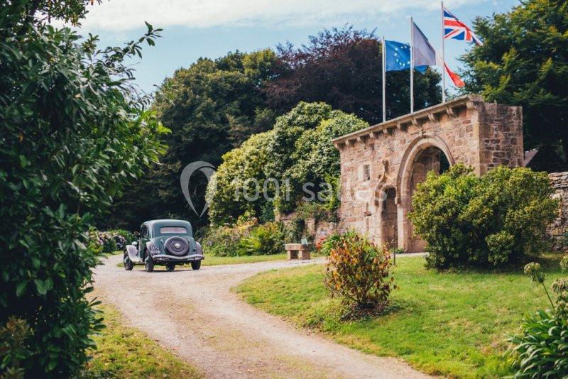Une voiture ancienne roule sur une allée bordée de verdure près d'un bâtiment en pierre avec des drapeaux.