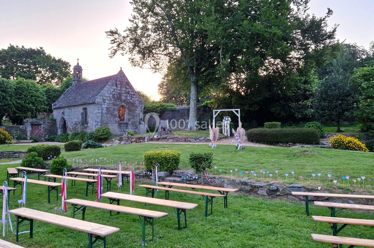 Bancs en bois disposés en extérieur devant une petite chapelle en pierre entourée de verdure au coucher du soleil.