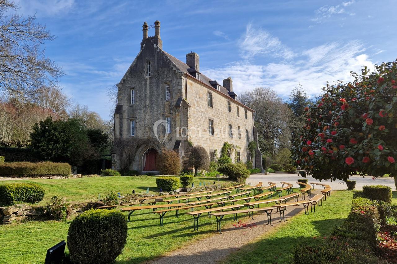 Grand bâtiment en pierre entouré de jardins, avec des bancs disposés en extérieur sous un ciel dégagé.