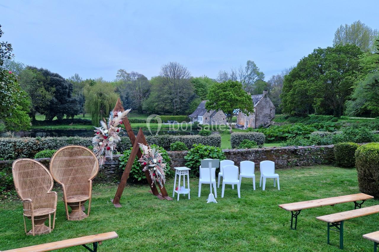 Décor de cérémonie en plein air avec chaises blanches, bancs en bois, arche florale et vue sur un jardin verdoyant.