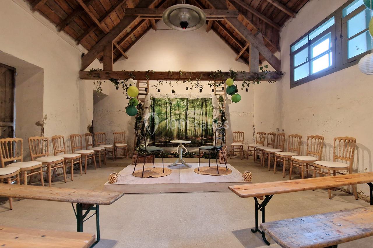 Salle rustique aménagée pour une cérémonie, avec chaises en bois, bancs et décor forestier en arrière-plan.