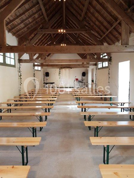 Salle avec charpente en bois, bancs alignés et scène équipée de micros et enceintes au fond.