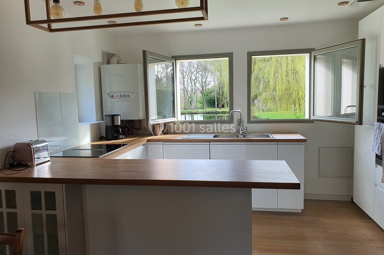 Cuisine lumineuse avec plan de travail en bois, fenêtres ouvertes donnant sur un jardin avec arbres et étang.