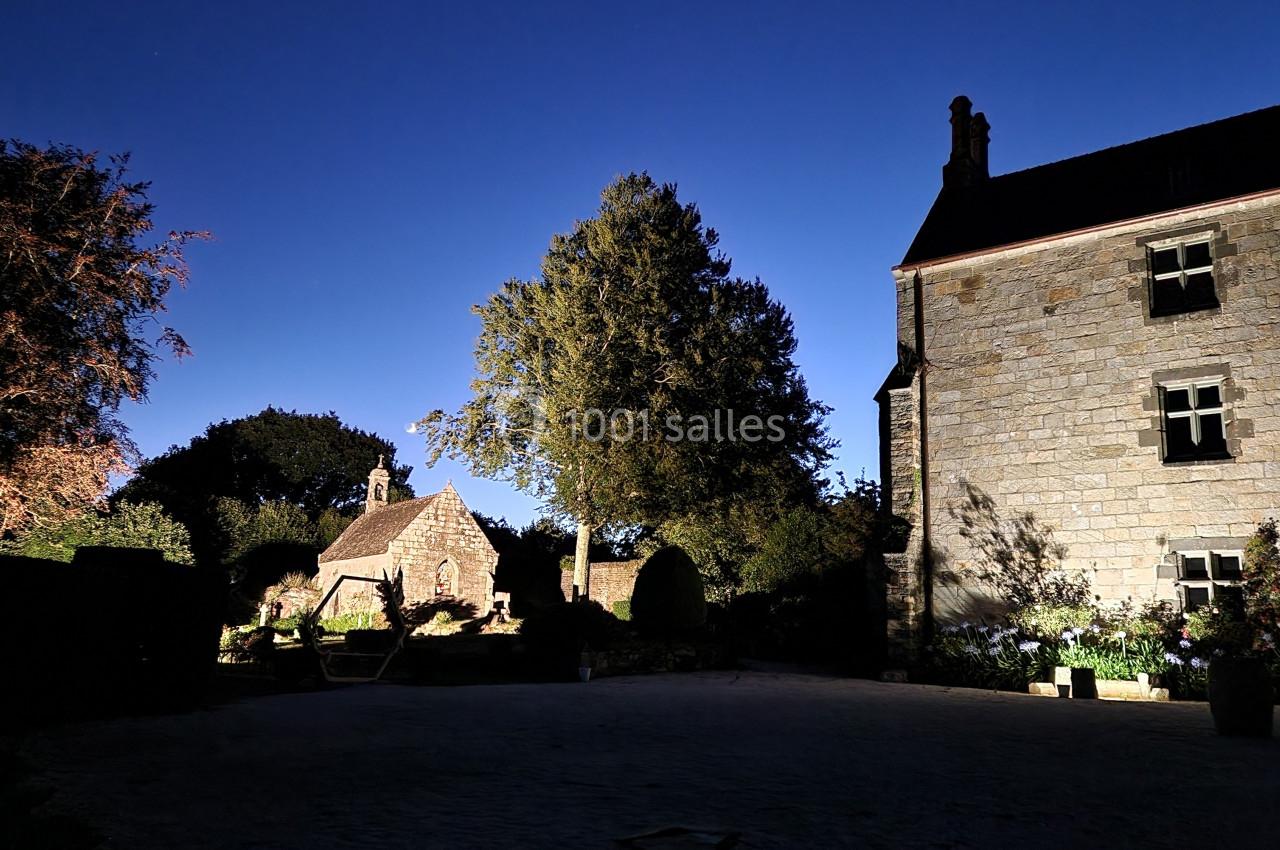 Cour d'une maison en pierre éclairée la nuit, avec un arbre central et une petite chapelle en arrière-plan.