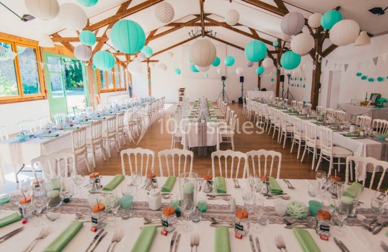 Salle de réception décorée pour un événement, avec tables alignées, nappes blanches et lanternes suspendues.