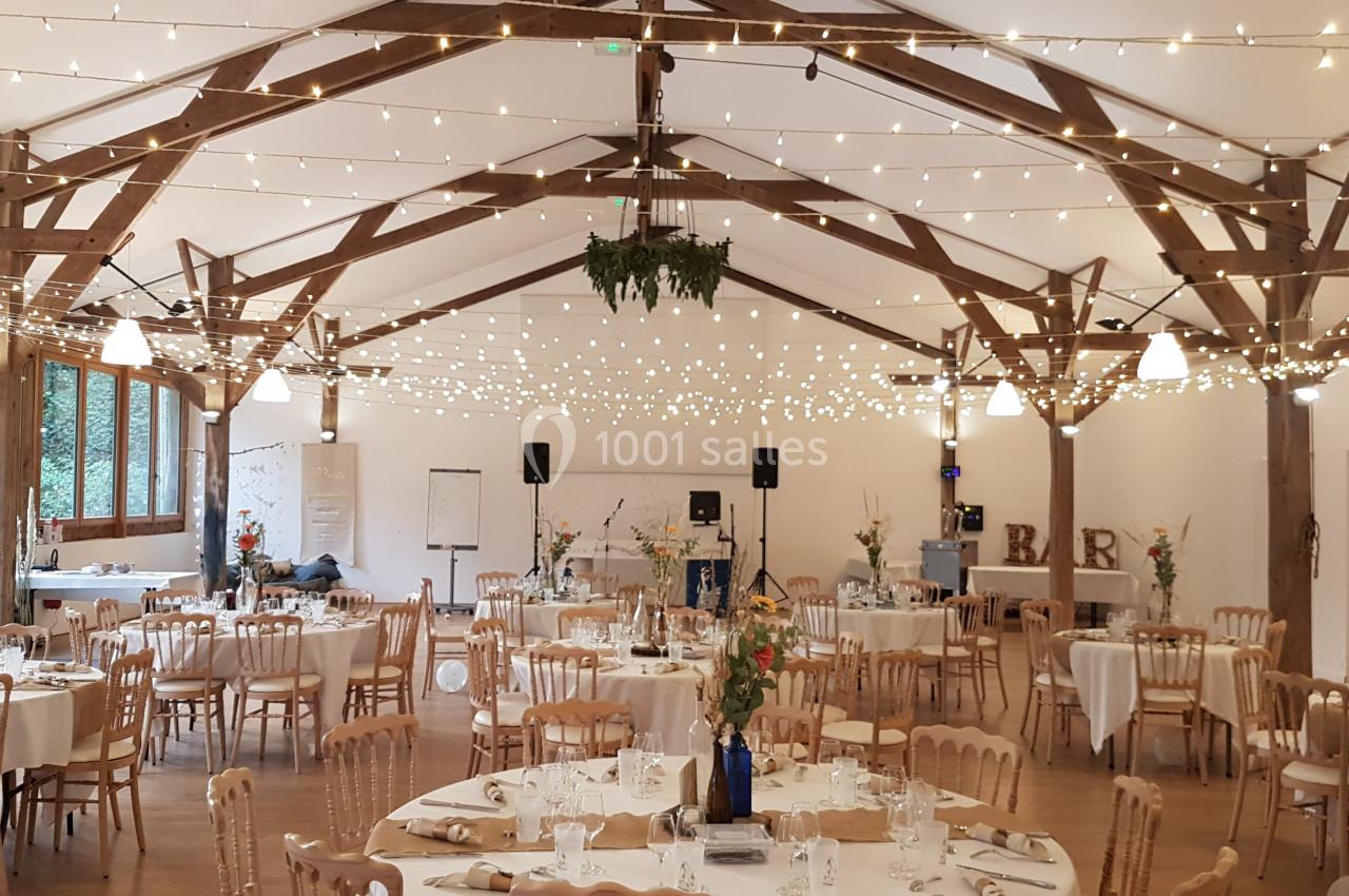 Salle de réception décorée avec des guirlandes lumineuses, des tables rondes dressées et des chaises en bois clair.