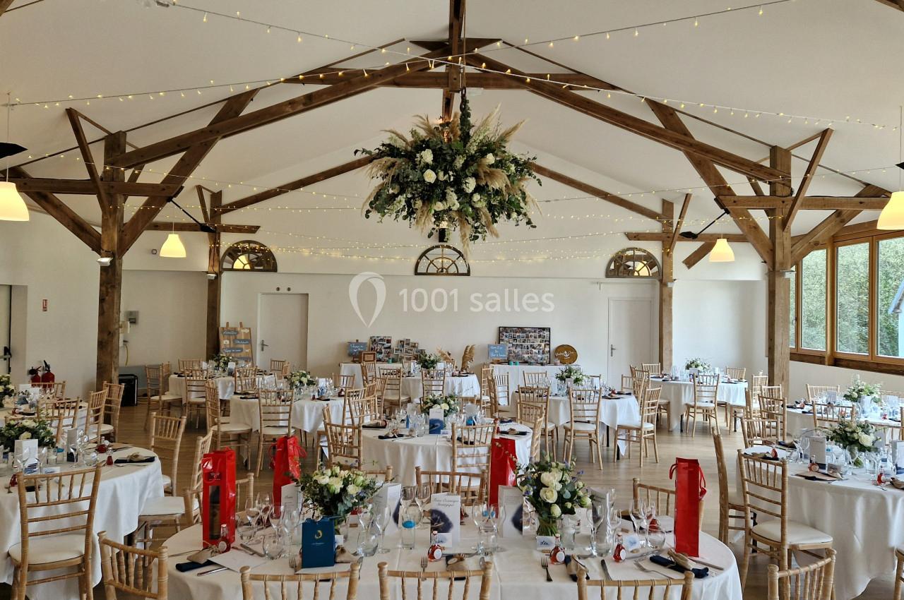 Salle de réception décorée avec des tables rondes, des chaises en bois, des fleurs et des guirlandes lumineuses suspendues.