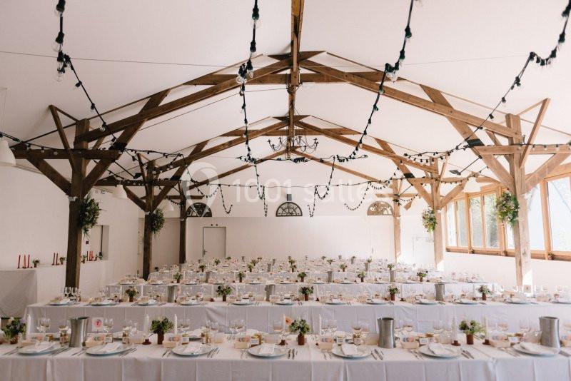 Salle de réception décorée avec des tables alignées, nappes blanches, guirlandes lumineuses et poutres apparentes en bois.