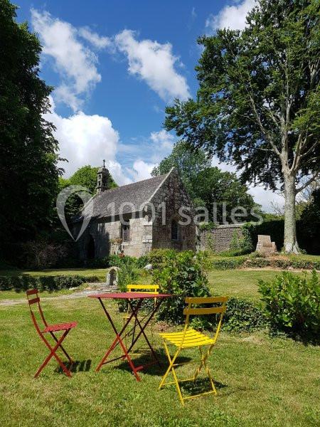 Petite chapelle en pierre entourée de verdure avec une table et des chaises colorées sur une pelouse ensoleillée.