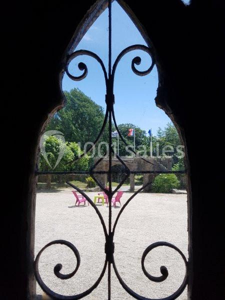 Vue d’un jardin avec des chaises roses et des drapeaux, encadrée par une fenêtre à grille en fer forgé.