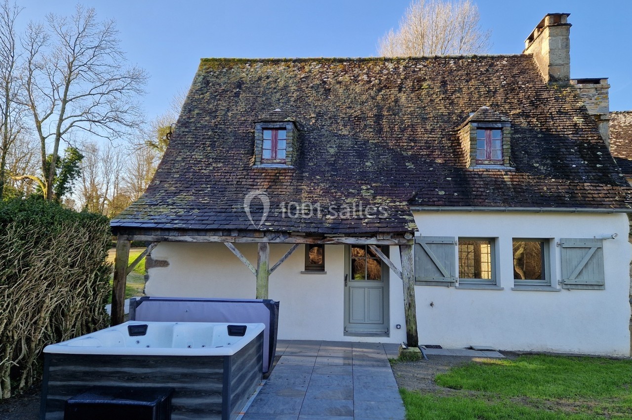 Façade d'une maison en pierre avec toit en ardoise, jacuzzi sur une terrasse en pierre et haie à gauche.