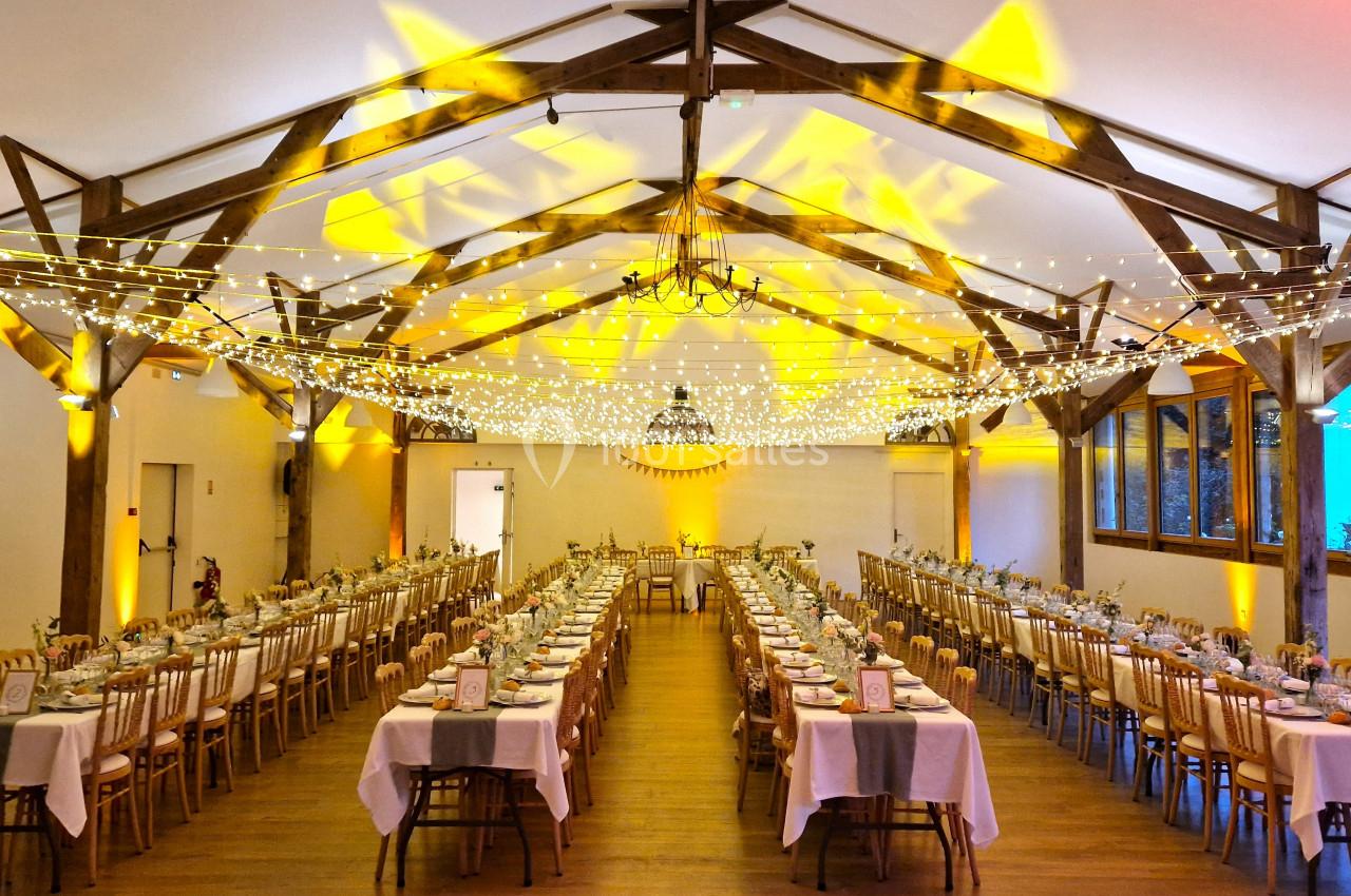 Grande salle de réception décorée avec des guirlandes lumineuses, des tables alignées et des chaises en bois.