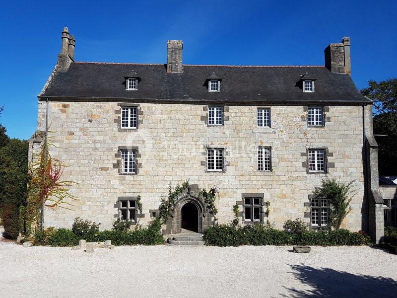 Façade d'une grande maison en pierre avec toit en ardoise, entourée de végétation et d'une cour gravillonnée.