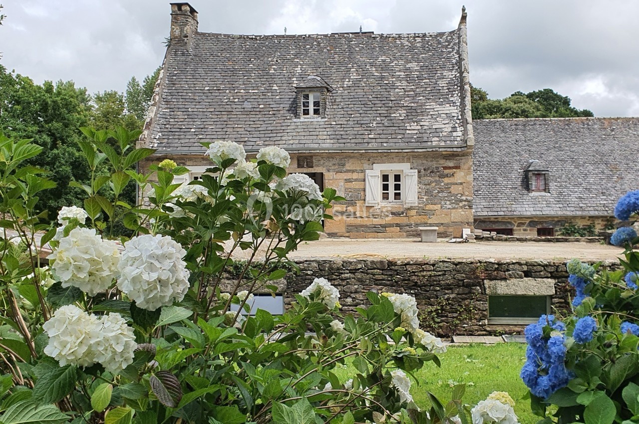 Maison en pierre traditionnelle avec toit en ardoise, entourée d'hortensias blancs et bleus dans un cadre verdoyant.