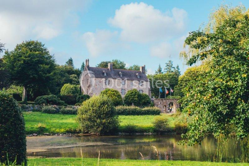 Manoir en pierre entouré de verdure, avec un étang au premier plan et un ciel partiellement nuageux.