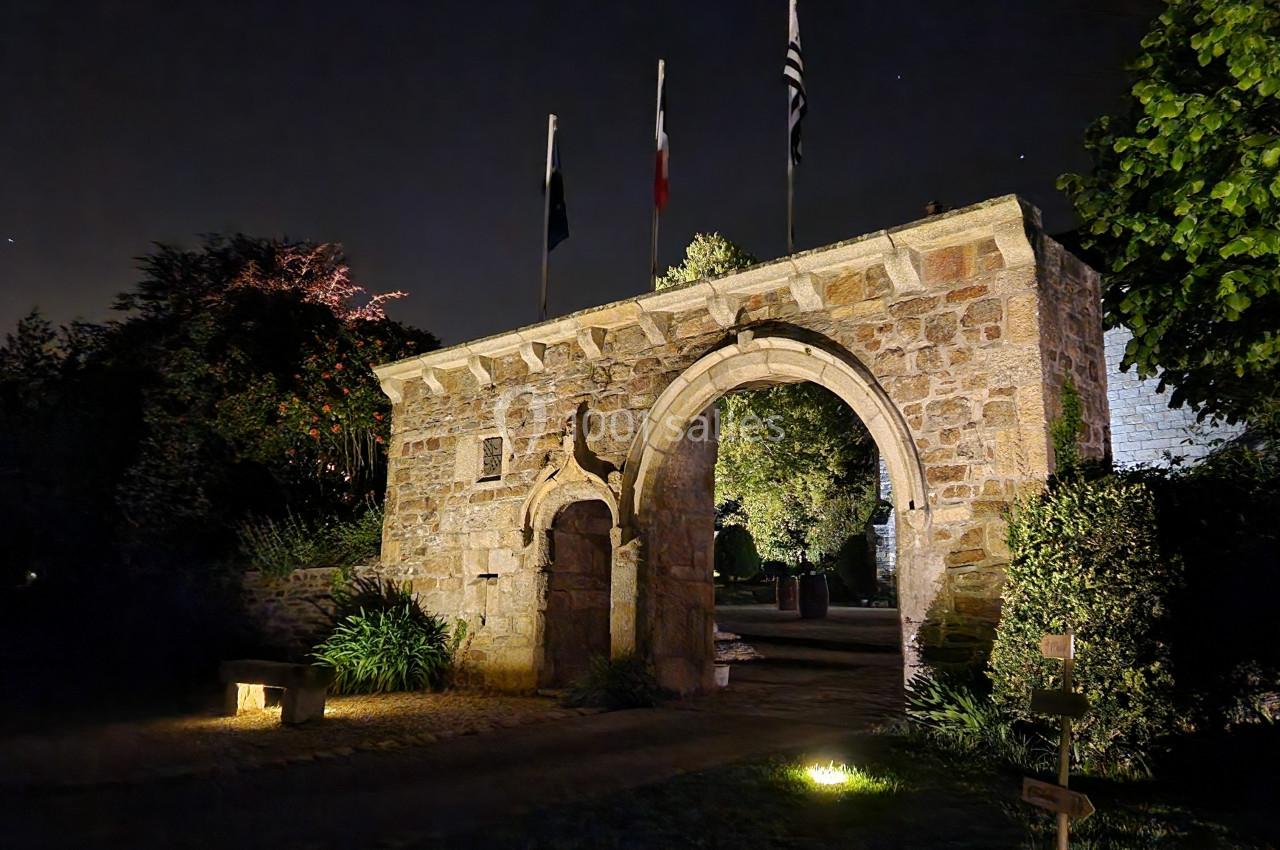 Entrée en pierre éclairée de nuit avec un arc voûté, des drapeaux au sommet et un jardin en arrière-plan.