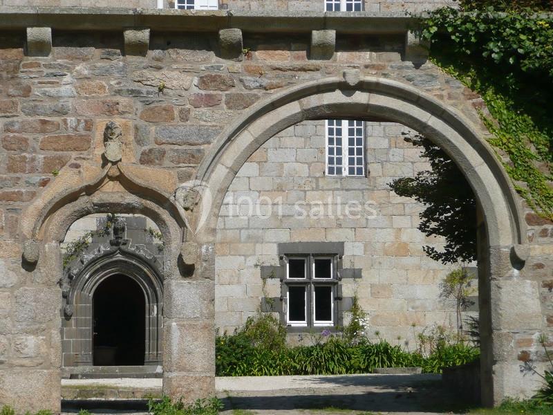 Arcades en pierre devant une façade en pierre avec fenêtres, entourées de végétation.