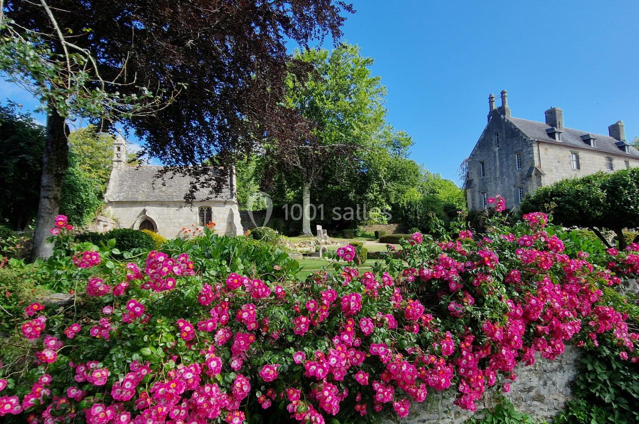 Jardin fleuri avec des roses roses au premier plan, église en pierre et bâtiment historique entourés de verdure en arrière…