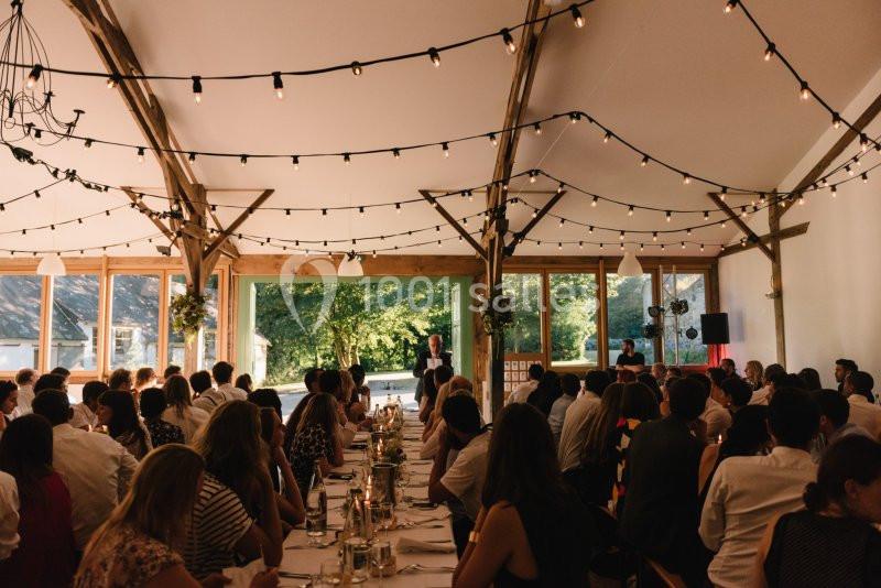 Salle de réception décorée avec des guirlandes lumineuses, remplie d'invités assis à des tables lors d'un événement.