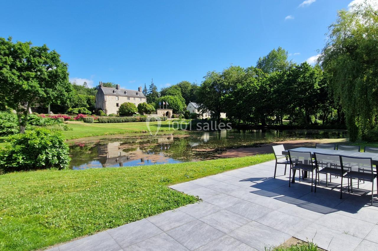 Terrasse avec table et chaises donnant sur un jardin verdoyant, un étang et une bâtisse en pierre sous un ciel bleu.