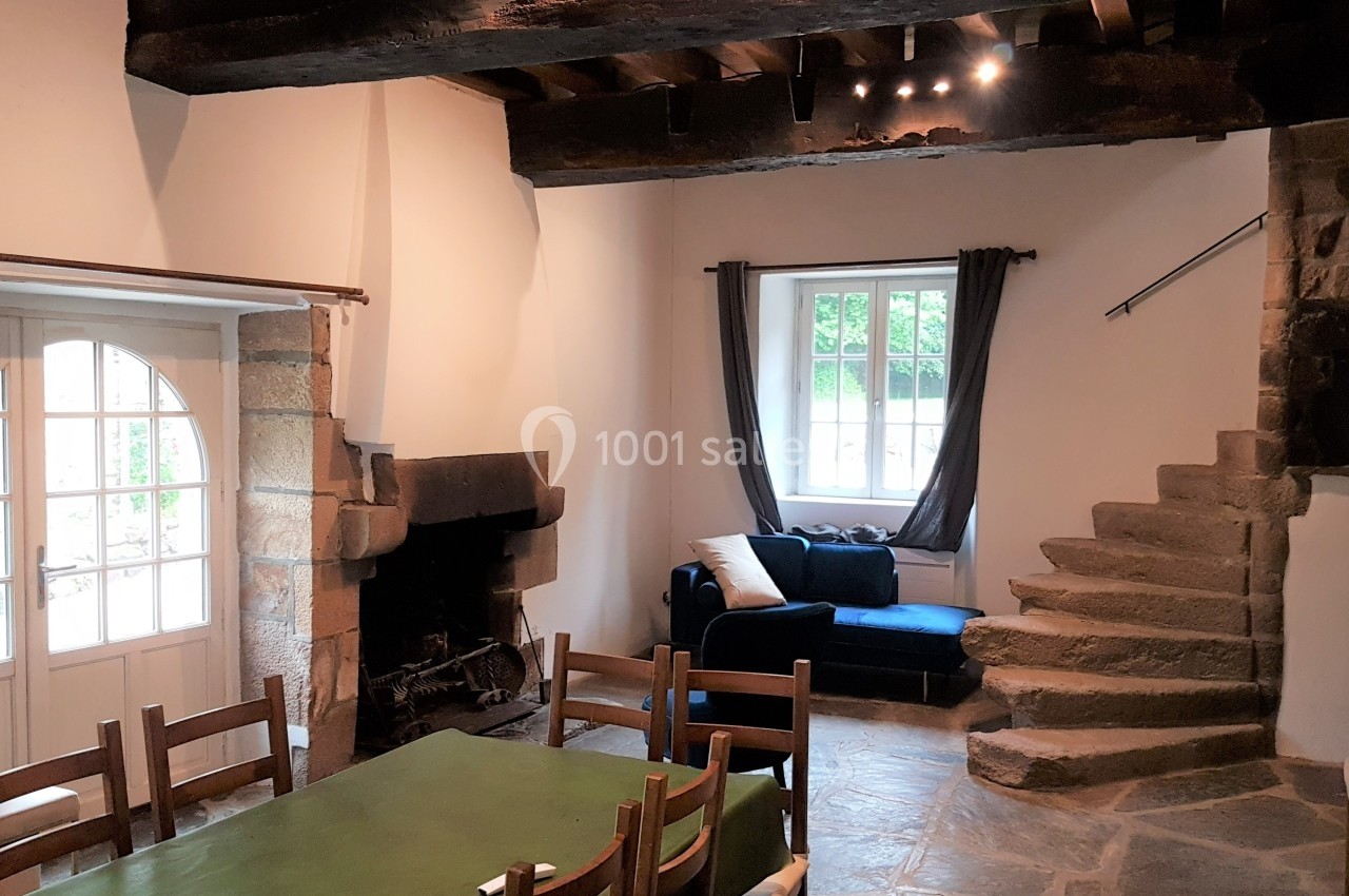 Salle de séjour avec cheminée en pierre, table en bois, canapé bleu, escalier en pierre et lumière naturelle.