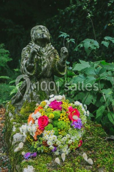 Statue en pierre d'une figure religieuse entourée de végétation, avec un bouquet de fleurs colorées au premier plan.