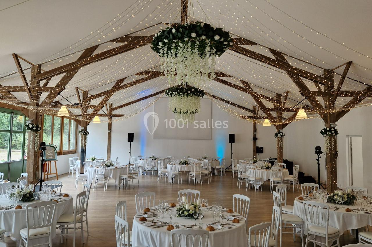 Salle de réception décorée avec des guirlandes lumineuses, tables rondes dressées et poutres apparentes en bois.