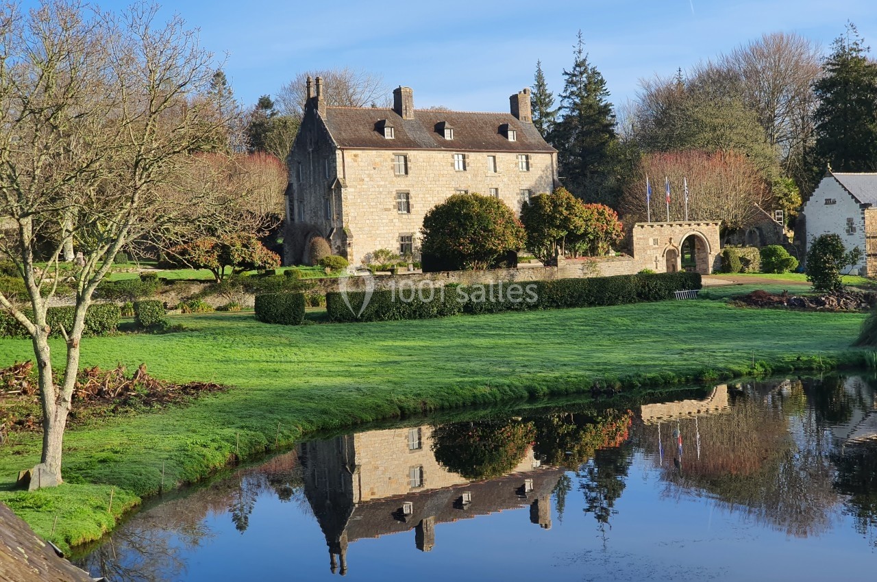 Manoir en pierre entouré de jardins verdoyants, avec un étang reflétant le bâtiment et le paysage environnant.