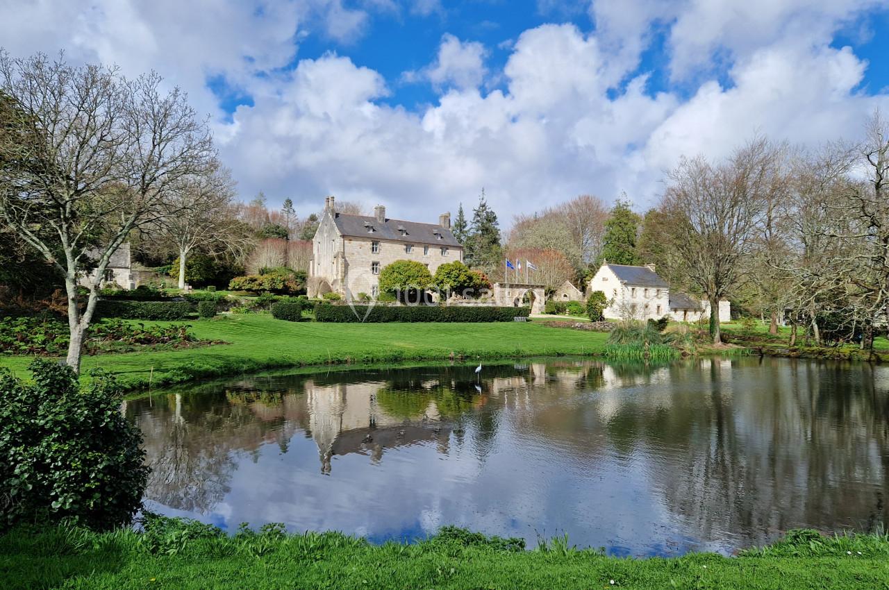 Vue d'un manoir en pierre entouré de jardins verdoyants, avec un étang reflétant le bâtiment et le ciel nuageux.
