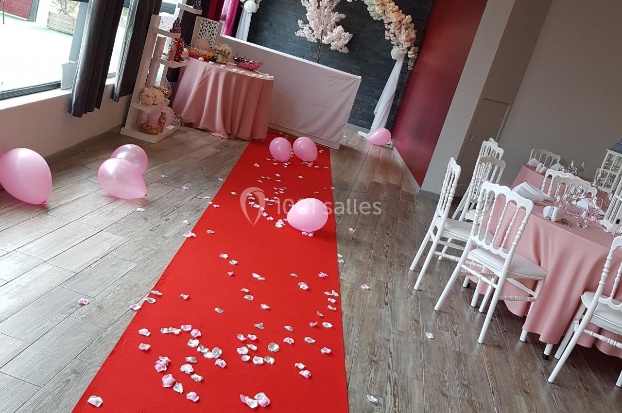 Salle décorée pour un événement avec tapis rouge, pétales, ballons roses et arche florale blanche en arrière-plan.