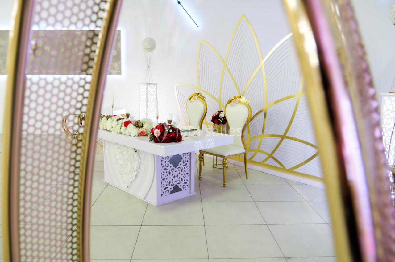 Salle décorée avec une table blanche ornée de fleurs rouges et blanches, deux chaises dorées et un motif mural doré.