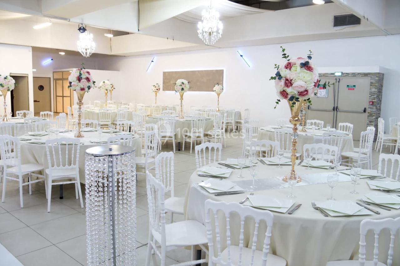 Salle de réception décorée avec des tables rondes, nappes blanches, chaises assorties et centres de table fleuris.