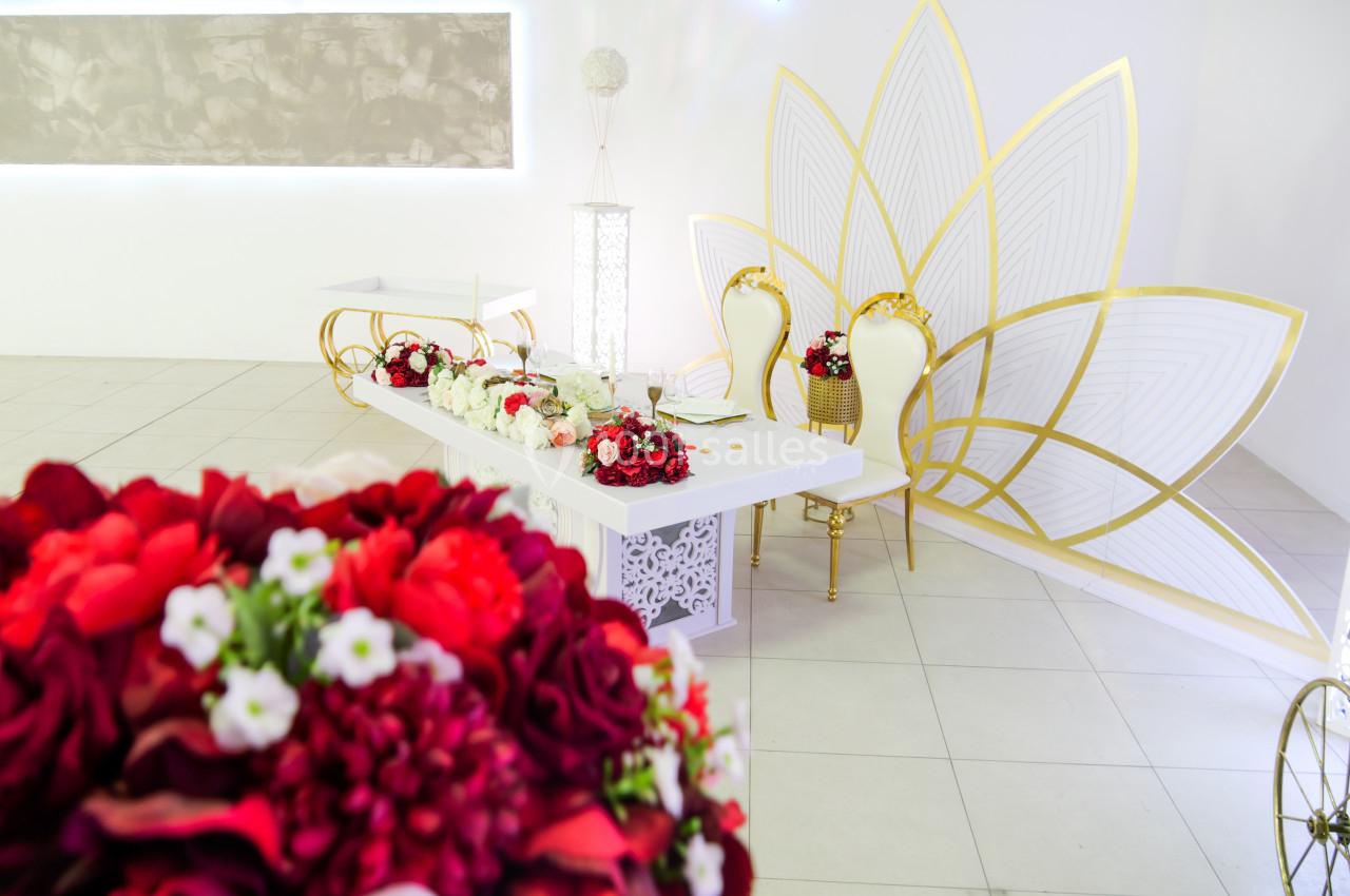 Salle décorée pour un événement avec table ornée de fleurs rouges, chaises dorées et grand motif floral doré en arrière-plan.