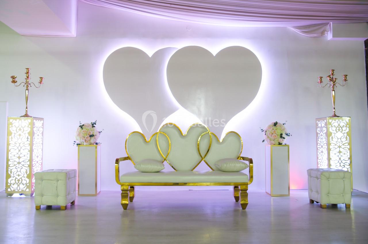 Canapé blanc et doré avec motifs de cœurs, entouré de décorations florales et éclairages doux dans une salle.