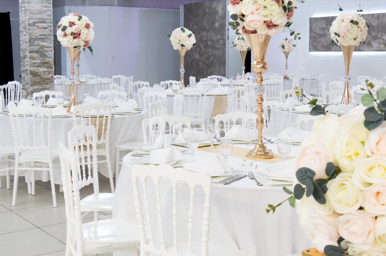 Salle de réception décorée avec des tables rondes, nappes blanches, chaises blanches et centres de table fleuris.
