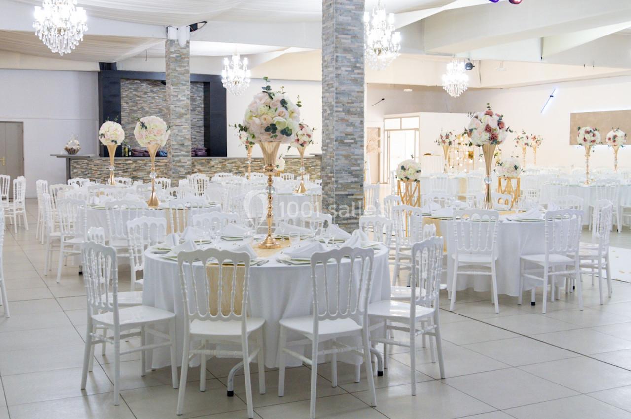 Salle de réception lumineuse avec tables rondes décorées de fleurs et chaises blanches, sous des lustres élégants.