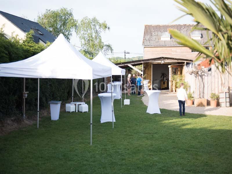 Jardin avec tentes blanches, tables hautes et quelques personnes près d'un bâtiment en bois.