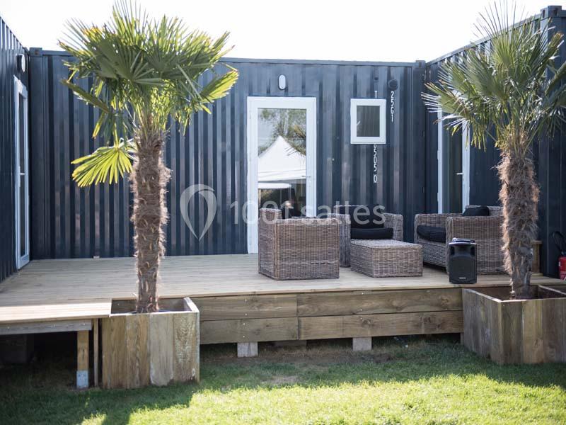 Terrasse en bois avec fauteuils en rotin, entourée de palmiers en pots, devant un bâtiment en conteneur noir.