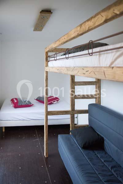 Lit superposé en bois au-dessus d'un canapé bleu, avec un lit double et des coussins rouges en arrière-plan.